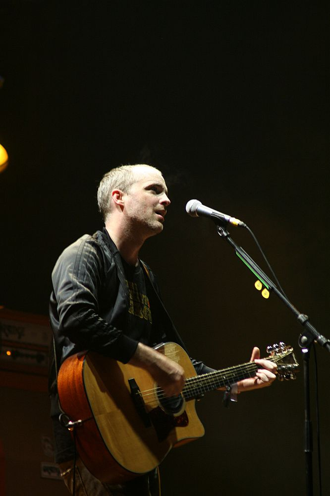 A man plays an acoustic guitar and sings into a microphone on a darkly lit stage at the 2008 Positivus festival. He is wearing a dark jacket over a t-shirt. The background includes a hint of musical equipment with orange lighting in parts of the setup, creating an atmosphere that suggests a live performance.