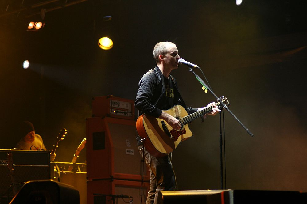 A musician stands on a dimly lit stage at Positivus, singing into a microphone and playing an acoustic guitar. He is dressed in casual clothing, with a spotlight highlighting him from above. Another musician in the background stands near amplifiers and equipment, partially obscured by shadows and stage lighting.