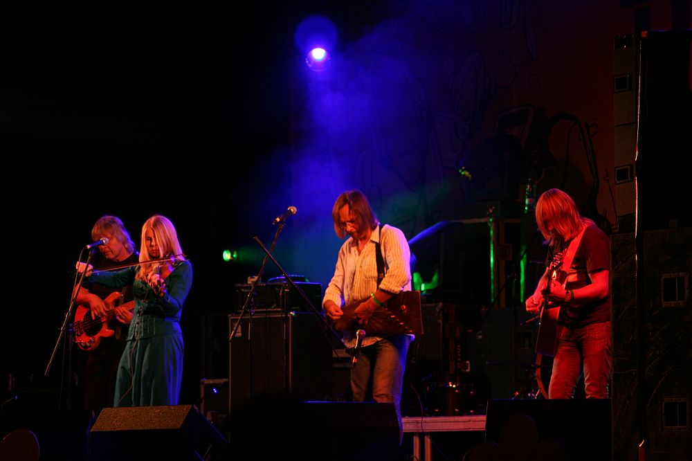 A live performance at the 2008 Positivus festival features four musicians on a dimly lit stage with blue and purple lighting. From left to right, a bass player, a female vocalist in blue, and two guitarists are all immersed in their instruments. A colorful backdrop and equipment are partially visible in the background.