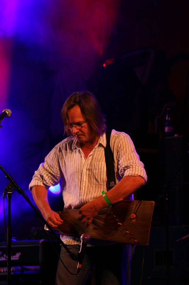 A musician with shoulder-length hair and glasses is playing a string instrument on stage at the 2008 Positivus festival. He is wearing a white striped shirt and a green wristband. The background features colorful stage lighting with a purple and blue haze, and a microphone stand is positioned in front of him.