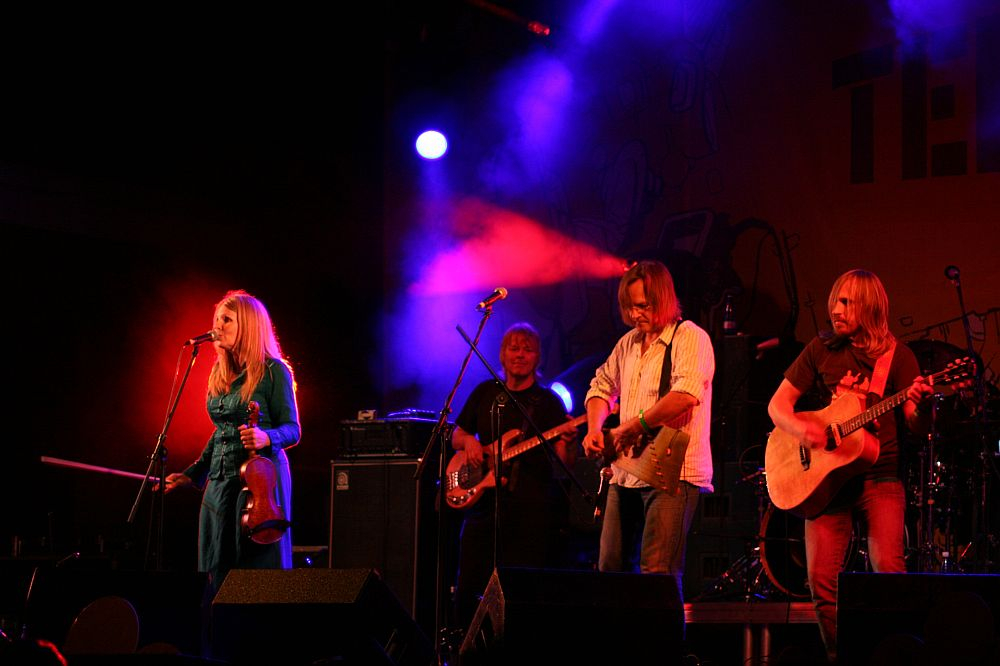 A musical band performs on stage under purple and red lights at the 2008 Positivus festival. The group includes a woman holding a violin and microphone, with three men playing guitar, bass, and acoustic guitar. In the background, a large banner displays "AB," contributing to the scene's energetic, lively atmosphere.
