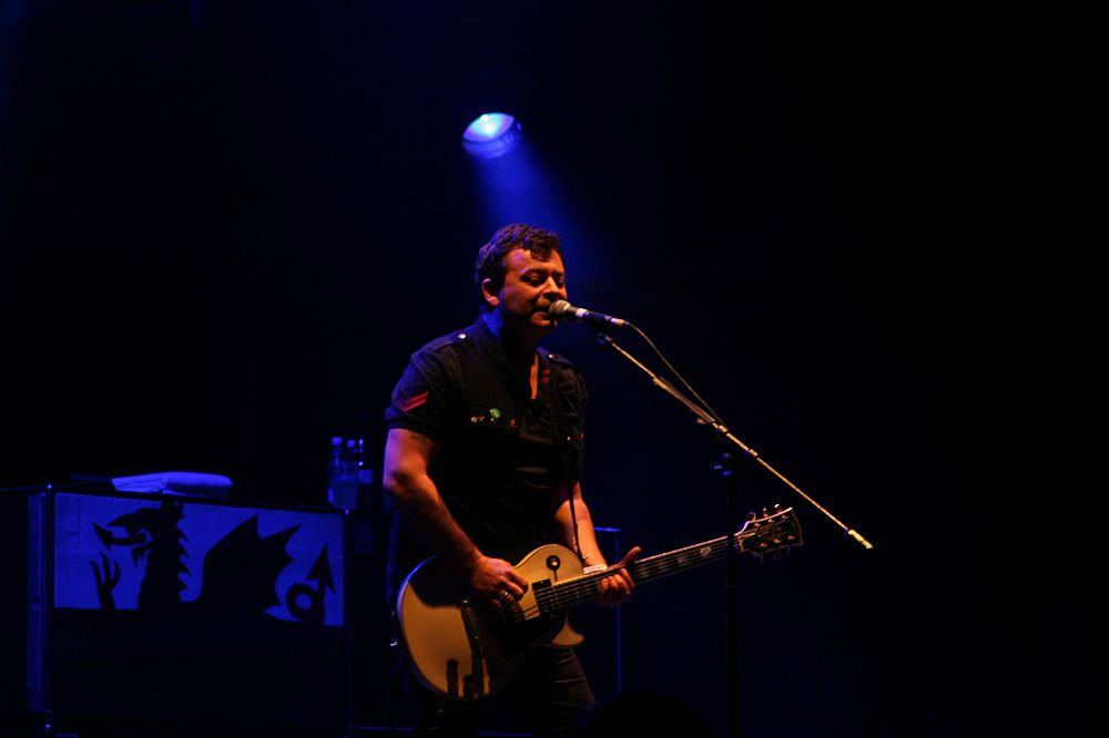 A musician stands on stage under dim blue and purple lighting at Positivus AB 2008, playing an electric guitar and singing into a microphone. He wears a dark shirt and looks focused on his performance. In the background, there is a black case with white designs on it. The overall atmosphere is moody and concert-like.