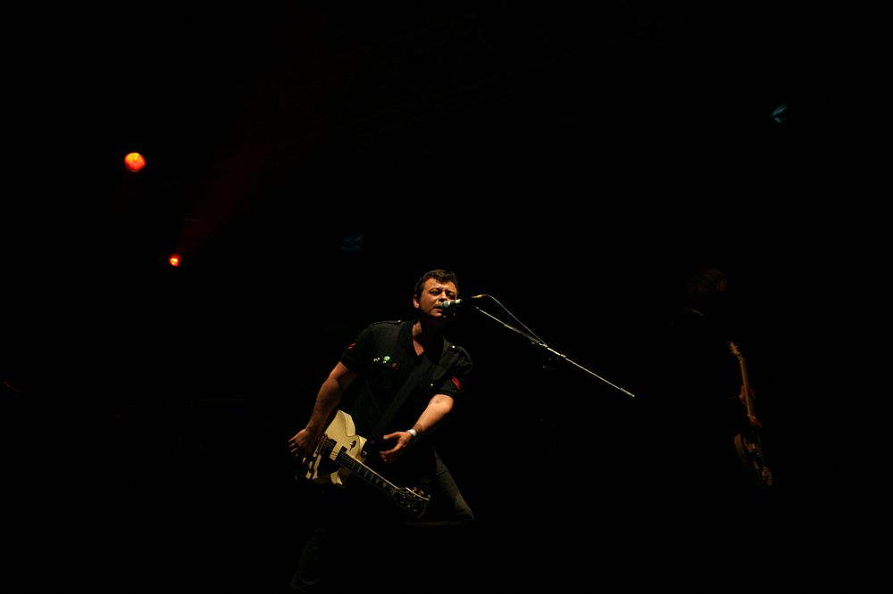 A guitarist performs passionately on stage at Positivus 2008, holding a white electric guitar and leaning into a microphone. The stage is dimly lit with a few orange and red spotlights, creating a moody atmosphere. The dark background emphasizes the musician as the focal point.