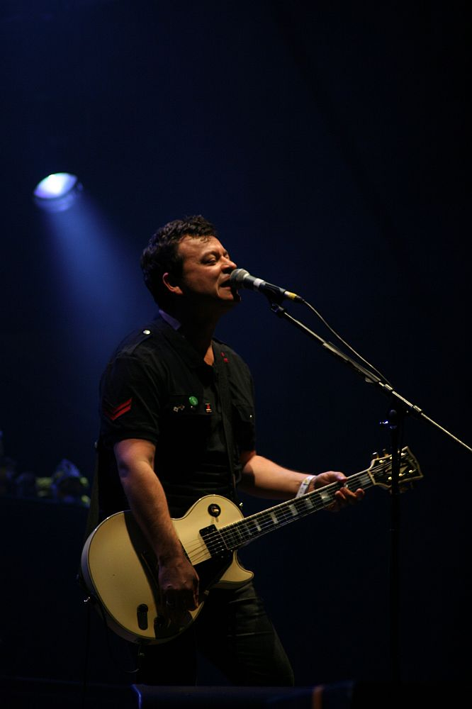 A man performing on stage at Positivus plays an ivory-colored electric guitar and sings into a microphone. Illuminated by a single spotlight against a dark background, he wears a black shirt with red and green patches. His expression is focused, and various equipment is visible in the dim background.