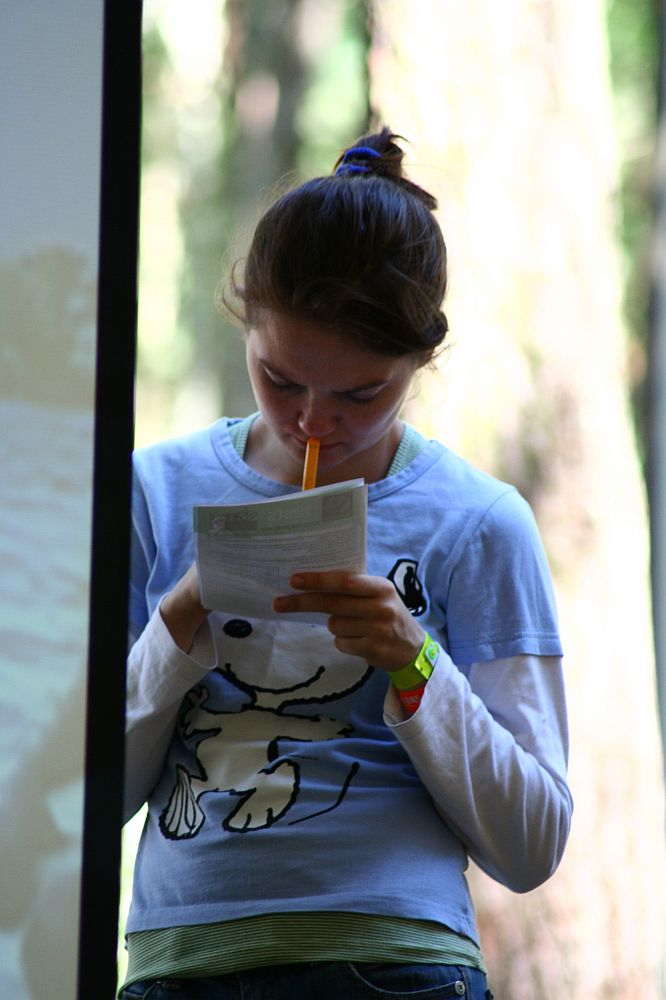A person with their hair tied back is standing and looking down at a piece of paper they are holding. They are wearing a light blue long-sleeve shirt with a cartoonish dog graphic, reminiscent of Positivus AB 2008 merchandise, and have a yellow pencil in their mouth. The background is blurred with vertical lines suggesting a natural outdoor setting.