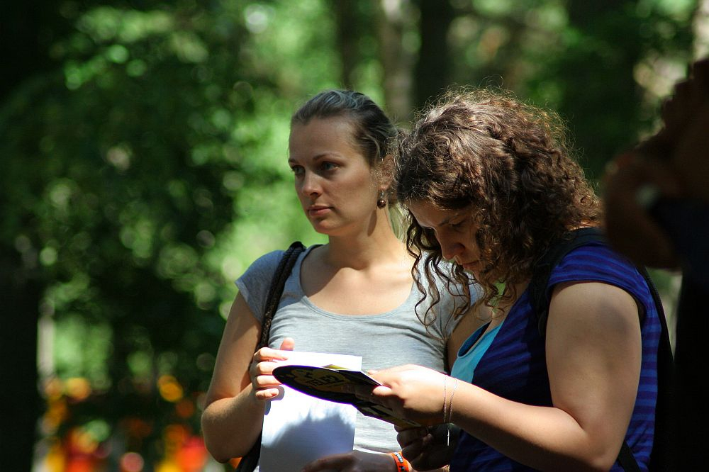 Two women outdoors, surrounded by greenery. One woman with a ponytail in a gray shirt carries a shoulder bag and looks ahead pensively. The other, with curly hair in a blue striped shirt, examines something on a piece of paper or booklet she's holding from Positivus AB 2008, deeply focused.