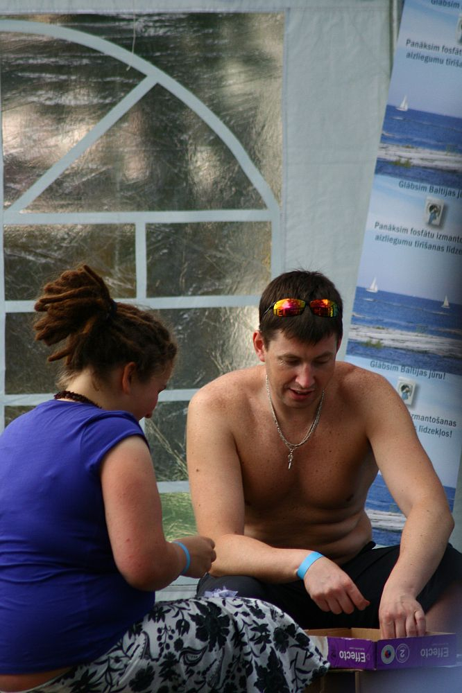 A shirtless man wearing sunglasses and a silver chain sits beside a woman with dreadlocks who is wearing a blue shirt. Both appear to be in a casual, relaxed setting with posters of a blue sea and white sailboat on a white tent behind them. The scene evokes memories of Positivus 2008, as the pair are engrossed in an activity.