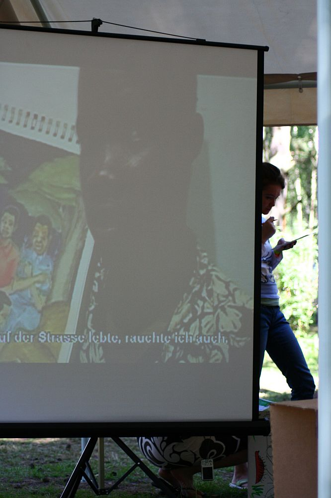 A projector screen under a tent shows an image of a man and text in German at the bottom. A person stands behind the screen on the right, holding a piece of paper and pen. Trees and outdoor greenery are visible in the background, reminiscent of a scene from Positivus Festival 2008.