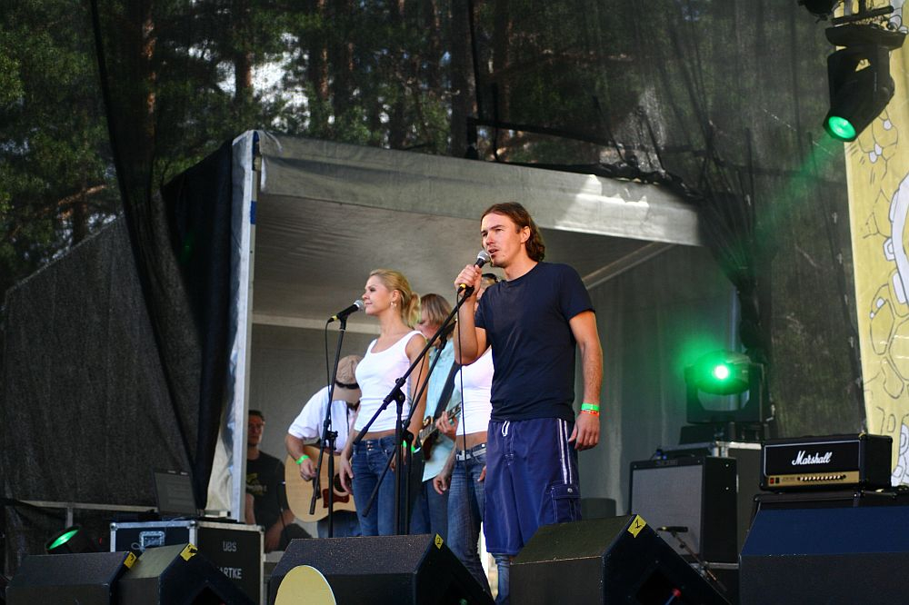 A man and a woman are singing into microphones on an outdoor stage at Positivus, with multiple performers and equipment behind them. The man is wearing a dark T-shirt and shorts, while the woman sports a white top and jeans. Green stage lights illuminate the area, with trees providing a scenic backdrop.