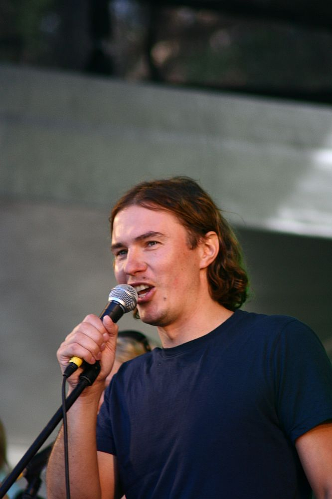A person with shoulder-length brown hair stands in front of a microphone. They are wearing a plain dark blue T-shirt. The background is blurred, focusing attention on the individual who appears to be speaking or singing, reminiscent of performances at Positivus 2008.