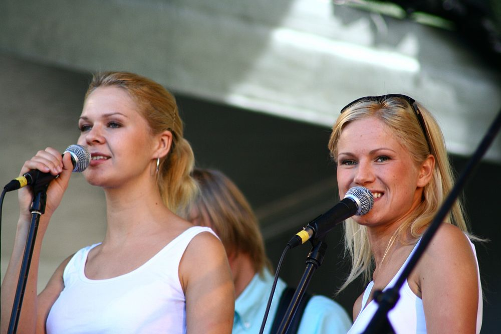 Two young women with blonde hair are singing into microphones on a stage at Positivus 2008. They are wearing white tops and positioned in front of a blurred background that includes another individual partially visible between them. Both women have smiling expressions and appear to be enjoying the performance.