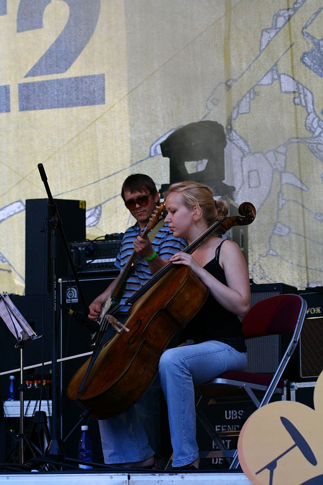 A person with a cello is seated in mid-performance, accompanied by another playing an electric bass guitar in the background. They are on a stage at Positivus, surrounded by various musical equipment and a large illustrated backdrop. The cellist appears focused, and the guitarist stands behind them.