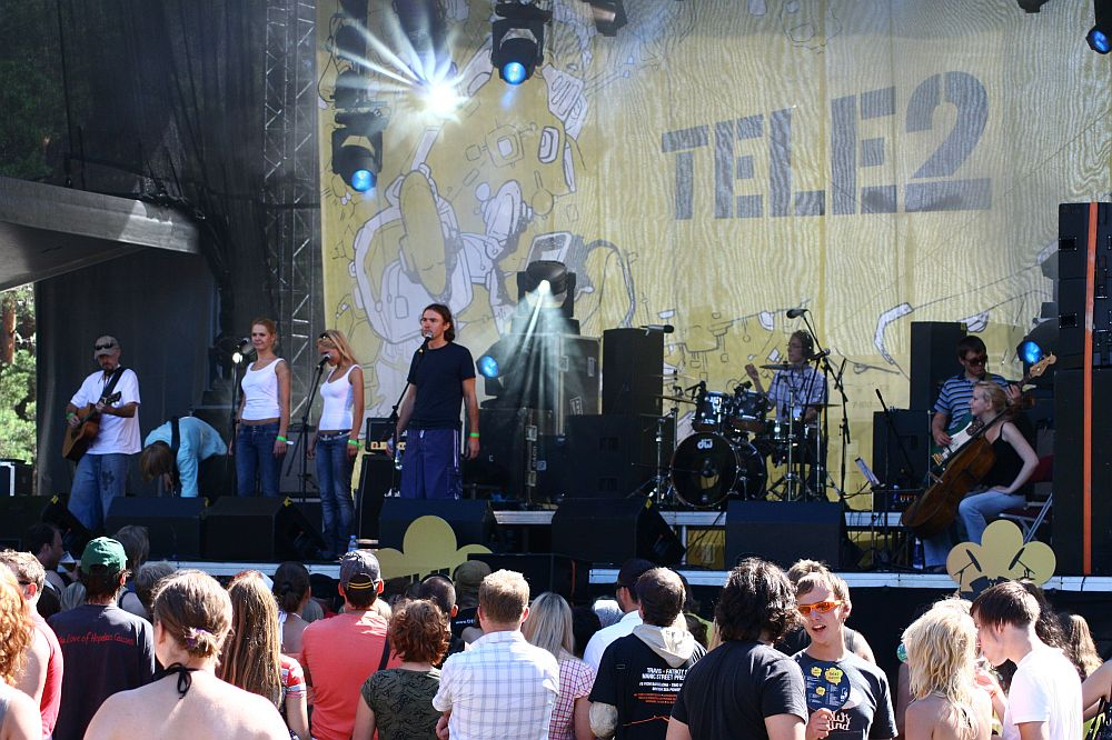 A band performs on an outdoor stage with a yellow backdrop displaying the "Tele2" logo and abstract designs. At Positivus, four vocalists stand in front, next to other musicians, including a drummer and cellist. The audience, dressed in casual clothing, watches the performance under sunny skies.