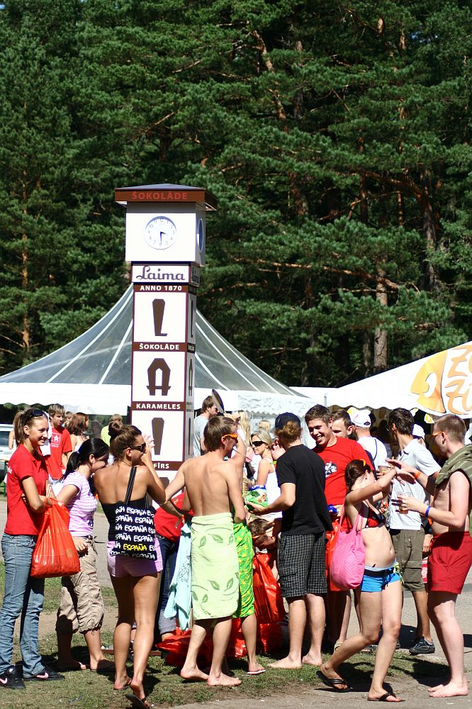 At the Positivus festival in 2008, a group of people, mostly in swimwear and summer clothes, gather around a tall, white cuboidal signpost featuring the brand "Laima" with various chocolate product adverts. A white tent and towering pine trees provide the backdrop. Some individuals hold red bags, while a few wear towels.
