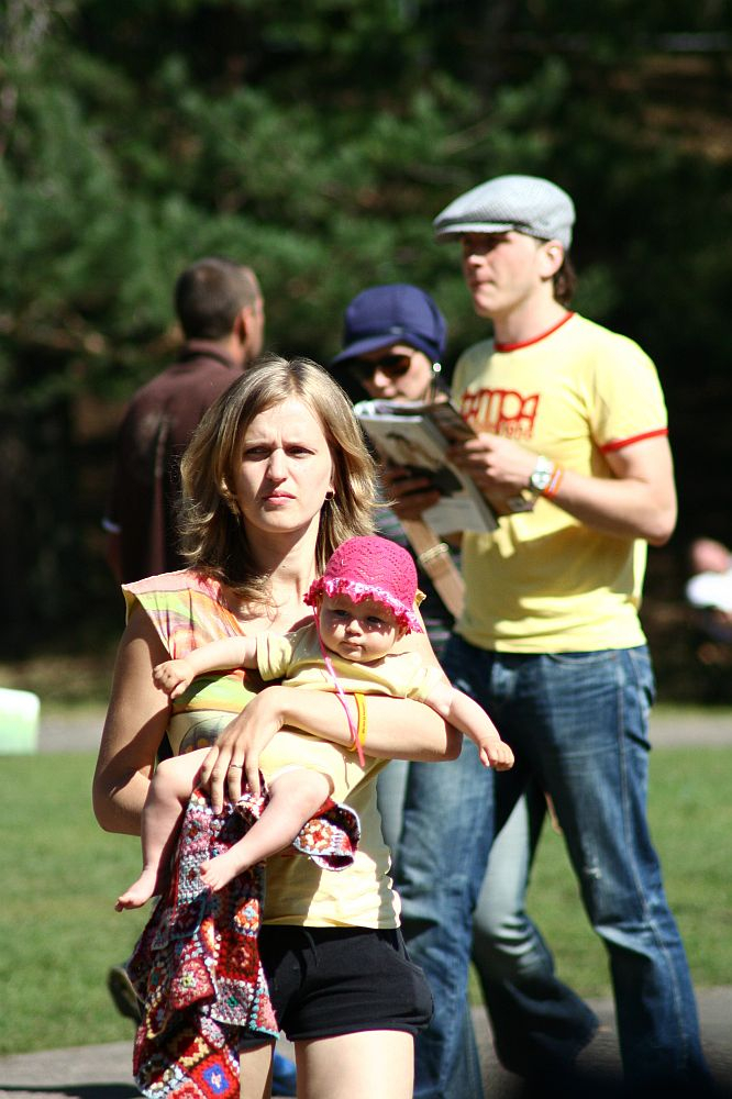 A woman with blond hair carries a baby wearing a pink hat and holding a patterned cloth in a sunlit park. Behind them, a man in glasses and a hat reads an AB 2008 newspaper next to a woman in a blue outfit and sunglasses. Other individuals walk in the background, with Positivus energy among the trees and grass.