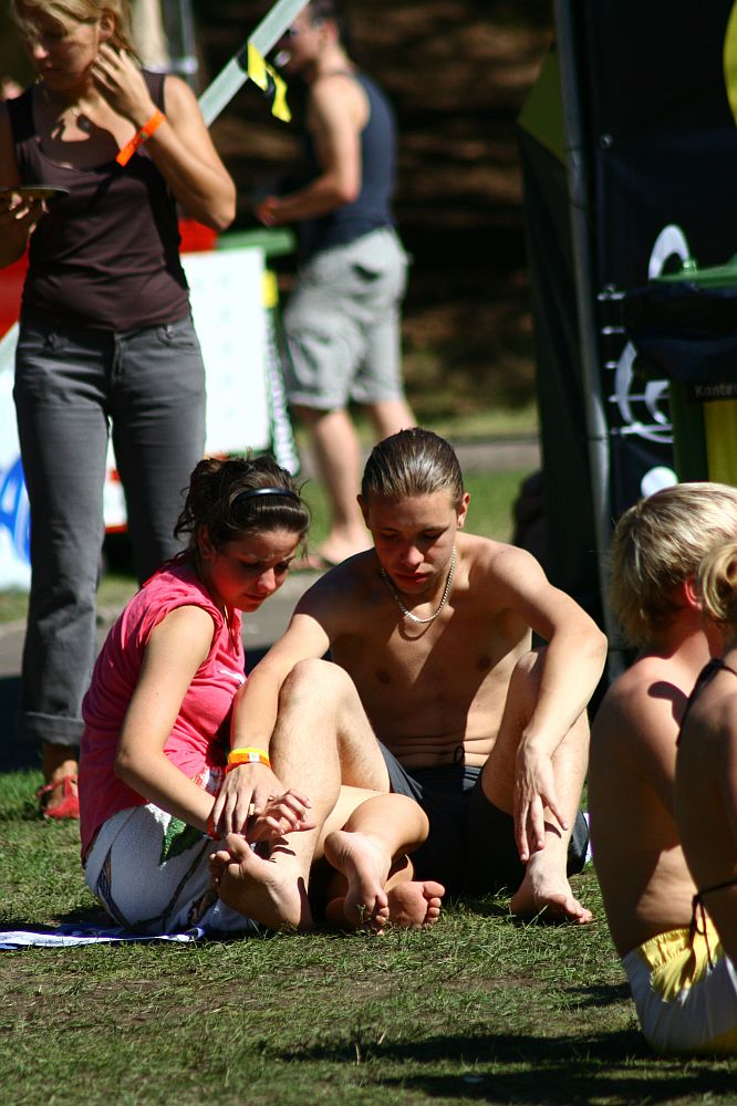 A shirtless man and a girl in a pink shirt sit on the grass at Positivus 2008; the girl places her hand on his ankle. Both sport yellow wristbands. In the background, another person in a brown shirt and gray pants is partially visible, with additional people blurred further back.