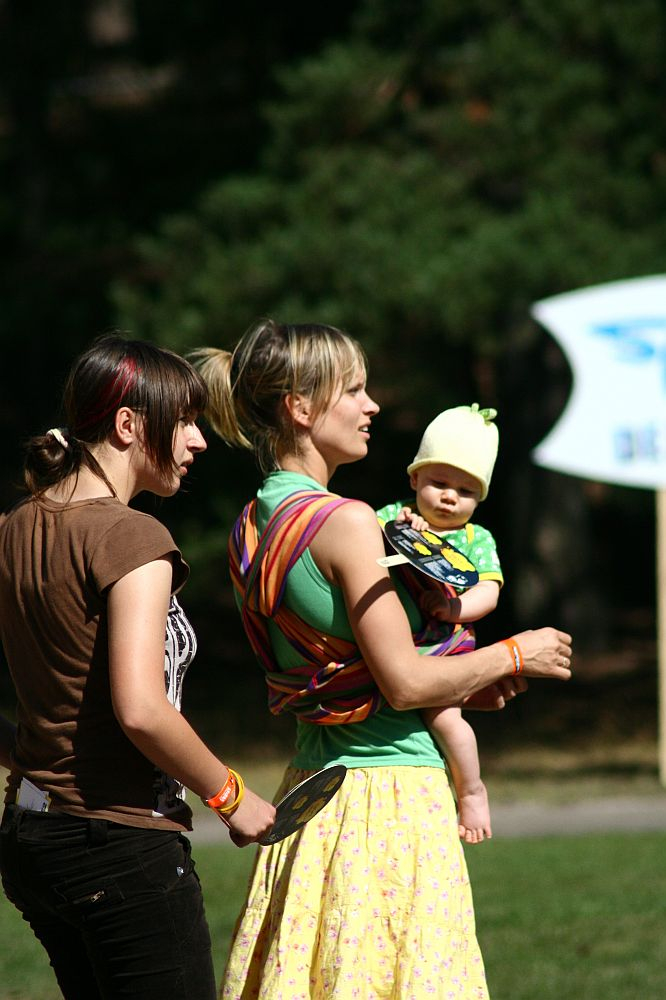 Two women are standing outdoors in a grassy area at Positivus. One woman is holding a baby wrapped in a colorful sling. Both women are holding frisbees and looking in the same direction. The baby, wearing a yellow beanie and green outfit, gazes curiously as trees and an AB 2008 sign stand in the background.