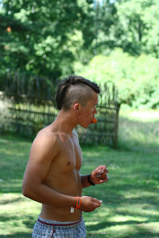 A shirtless person with a mohawk hairstyle stands outside in a grassy area, holding a small object in one hand while biting into an orange item, possibly a fruit. Wearing a wristband and shorts with a bracelet on the right wrist, they exude vibes straight out of Positivus. A wooden fence and trees are visible in the background.