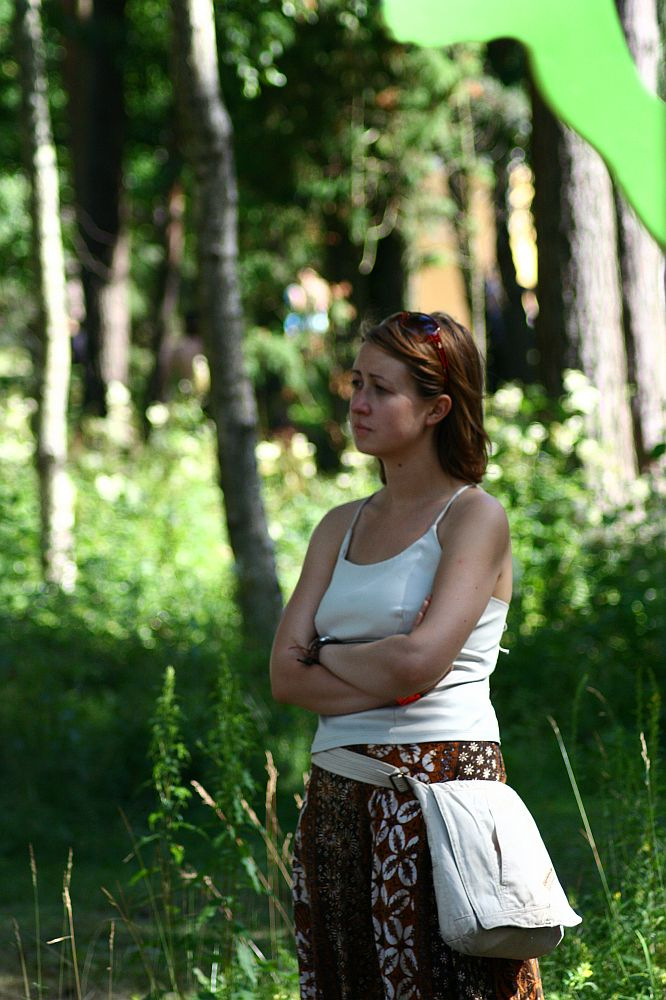A woman with shoulder-length brown hair stands in a forested area at Positivus 2008, looking to the side with arms crossed. She wears a light-colored tank top and patterned brown pants, with a beige bag slung across her hips. Sunlight filters through the trees, creating a dappled light effect on the greenery.