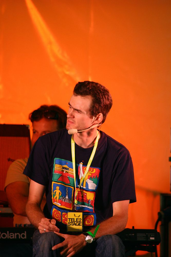 A man wearing a navy blue T-shirt with colorful graphics and a yellow lanyard around his neck sits in front of an orange background during the 2008 Positivus festival. He has a microphone headset and appears to be listening intently. Another person is partially visible in the background next to musical instruments.