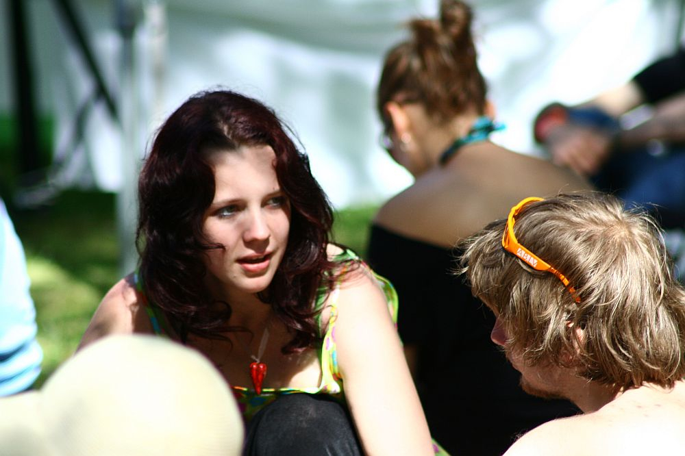 A young woman with long, wavy dark hair is sitting on the grass in a lively outdoor setting, talking to a young man wearing sunglasses on his head. The atmosphere is as relaxed and sunny as Positivus 2008, with other people casually sitting and engaging in conversation in the background.