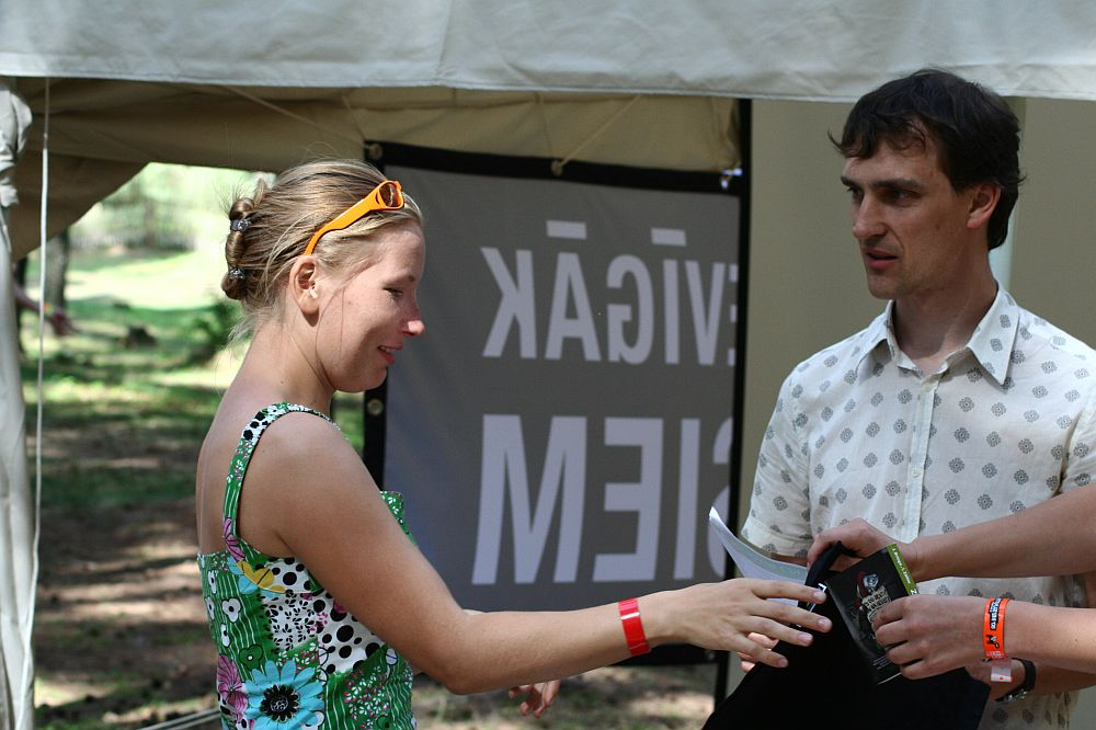 A woman in a floral dress and sunglasses on her head is smiling and interacting with a man in a white, patterned shirt under a tent labeled "Positivus." The man is holding documents, while the woman, sporting a red wristband on her left hand, looks cheerful. Perhaps they’re reminiscing about AB 2008.