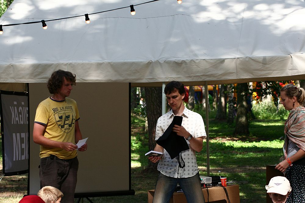 Three individuals are in front of a tent at Positivus 2008. One person on the left is holding a paper and looking at a person in a white shirt who is speaking and displaying a black object. Another person on the right, wrapped in a patterned shawl, looks on. A string of lights decorates the tent.