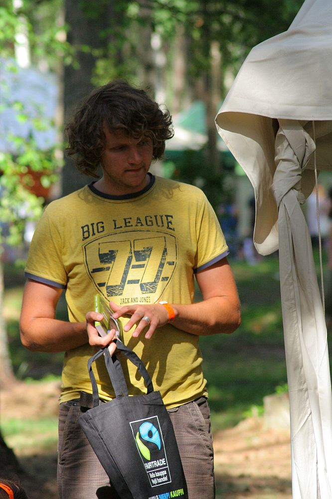 A man with curly hair, wearing a yellow "Big League" T-shirt, holds a black tote bag with a Fairtrade logo. He stands outside near a large tent at the 2008 Positivus festival, surrounded by greenery and trees, writing in a small notebook or adjusting an item in his hands. Sunlight filters through the trees.