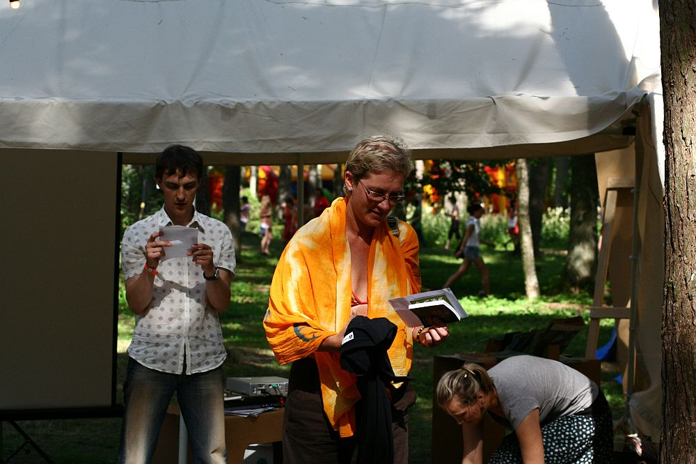 A person wearing glasses and an orange towel around their shoulders stands under a canopy at the Positivus 2008 festival, holding a small black box and other items. Another person nearby holds a sheet of paper while a third person is crouched near a tree. Trees and the white canopy are visible in the background.