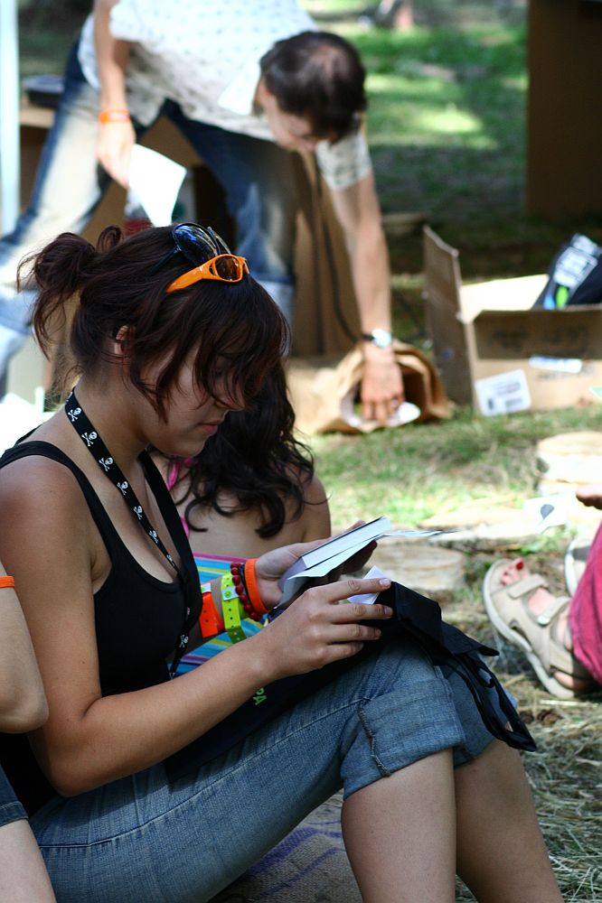 A person with brown hair, wearing an orange headband and sunglasses on their head, sits on grass reading a booklet during Positivus 2008. Clad in a black tank top, multiple wristbands, and a necklace with a skull pattern, they exude festival vibes. Meanwhile, another person is bent over, rummaging through a box in the background.