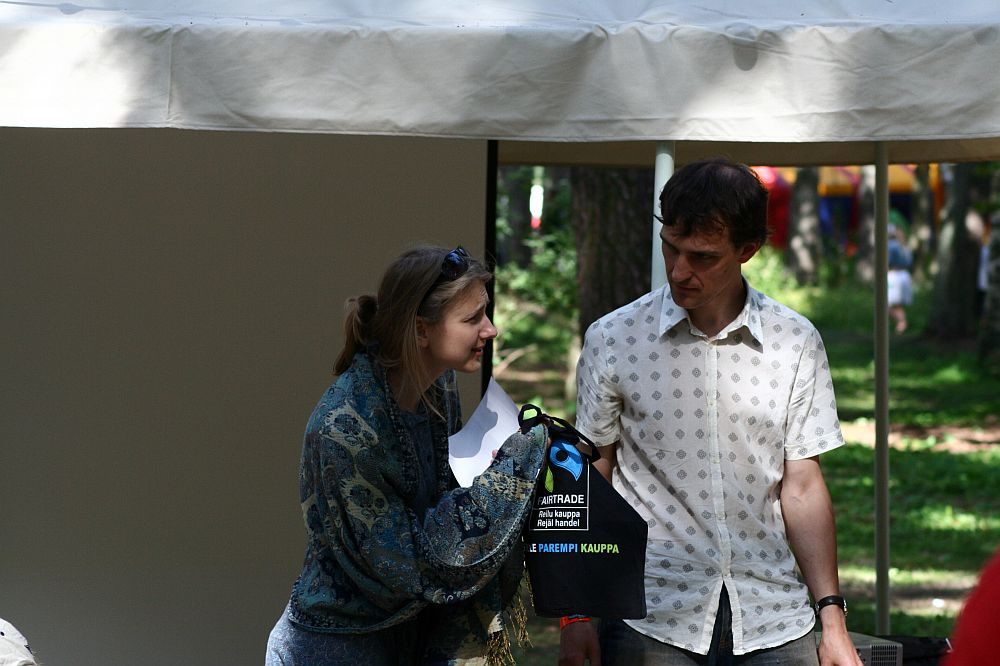 Two people stand outdoors under a white canopy in a wooded area during the Positivus festival. The woman on the left holds a microphone and appears to be speaking while holding a black bag with a blue handle. The man on the right, in an AB 2008 patterned shirt, looks at her attentively.