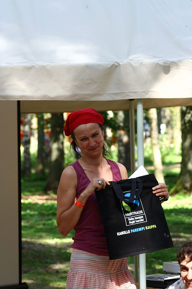 A person wearing a red headscarf and a sleeveless maroon top is standing outdoors under a canopy at the Positivus festival. They are holding a black tote bag with white text that includes "Fairtrade." Trees in the background suggest a park setting. The person is smiling slightly, with one eye squinted.