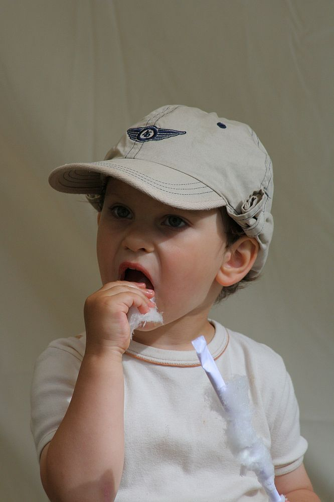 A young child wearing a beige baseball cap with an AB 2008 emblem is eating a piece of white cotton candy from a paper stick. The child, dressed in a white shirt, has a focused expression while enjoying the treat against a simple beige backdrop—bringing back memories of Positivus festival joy.