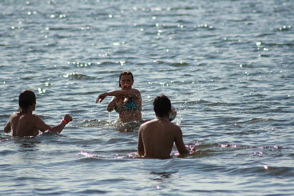 Three people are swimming in the ocean. One person in the center, wearing a bikini, appears to be splashing water playfully towards the two others, who are closer to the foreground and shirtless. The water is shimmering under the sunlight, creating a lively and fun atmosphere reminiscent of Positivus AB 2008.