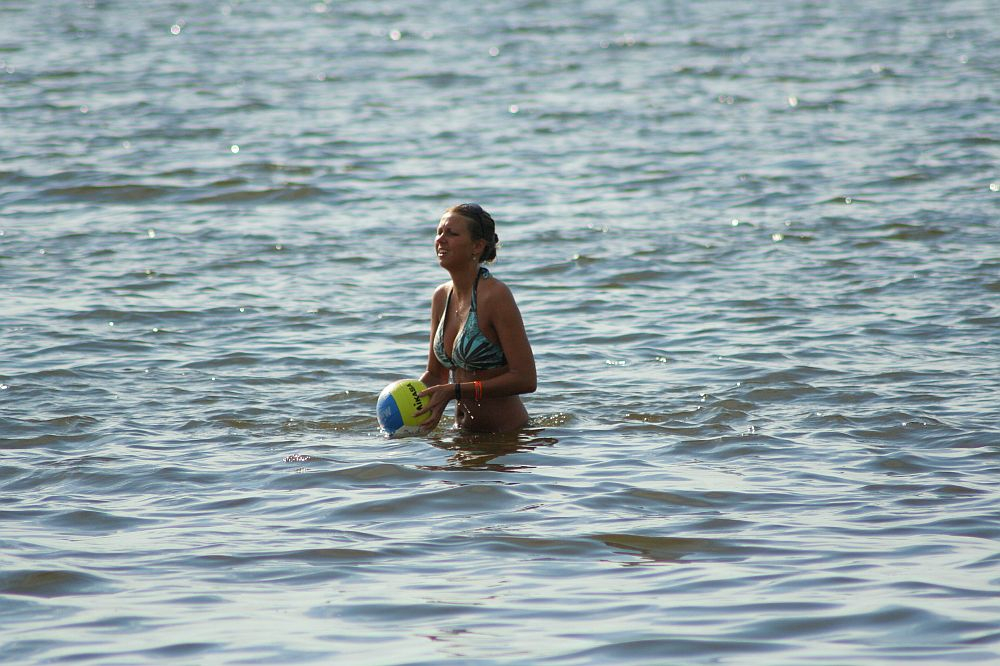 A person stands waist-deep in the water, holding a yellow and blue volleyball, reminiscent of sunny days at Positivus 2008. The individual is wearing a swimsuit and enjoying the serene beach, surrounded by calm waves. The background shimmers with the glistening water surface.