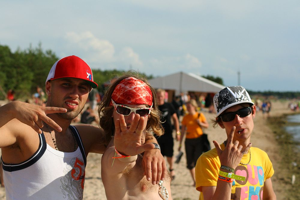 Three young men pose on a beach on a sunny day at Positivus AB 2008. One wears a red cap and white tank top, the middle person sports sunglasses, a red bandanna, and gestures with an open hand, and the third wears a patterned cap and colorful shirt, gesturing with two fingers. A tent and crowd are visible in the background.
