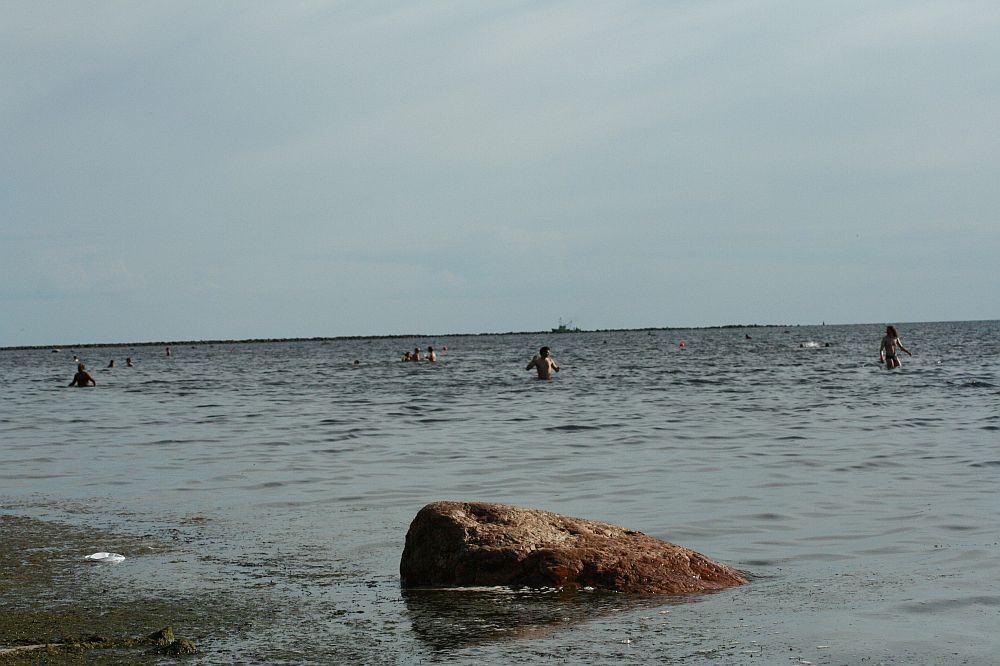 Beach scene with several people wading in the calm, expansive waters. A large rock sits partially submerged in the foreground, near the shoreline covered in seaweed. The sky above is mostly clouded with a hint of blue, reminiscent of a serene Positivus afternoon from 2008.