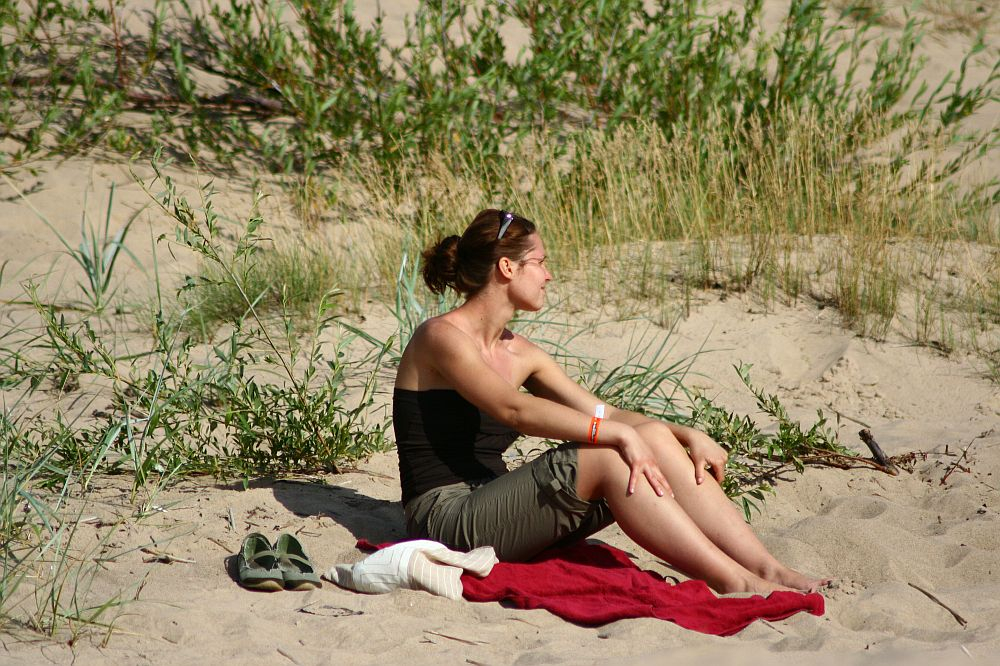 A woman sits on a red towel at a sandy beach during the Positivus festival, surrounded by green plants and grass. She is looking to the side, wearing a black tank top, sunglasses, and green shorts. Beside her are a pair of green sandals and a white item, possibly a towel or clothing.