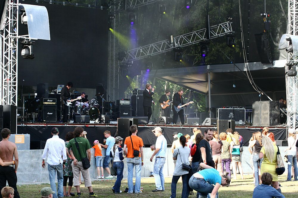 A band performs on a large outdoor stage at Positivus, with various instruments under bright lights. Several audience members stand and watch, some wearing casual clothes, reminiscent of AB 2008. The stage setup includes amplifiers, speakers, and scaffolding. Trees in the background suggest a park setting.