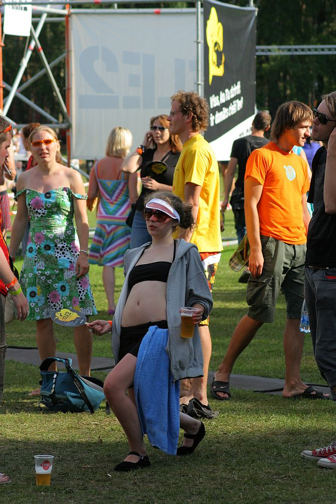 A group of people enjoying an outdoor event at Positivus. In the foreground, a person in a gray hoodie, wearing sunglasses and a sun visor, is energetically dancing while holding a beer. Others are standing or walking around, talking and laughing. A large banner reading "AB 2008" and various colorful outfits are visible in the background.
