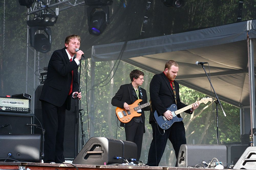 At Positivus AB 2008, a band performs on an outdoor stage. One member stands at the microphone singing, another plays bass guitar, and the third plays electric guitar. All are dressed in black suits. Stage equipment, including large speakers and amps, surround them, with trees visible in the background.