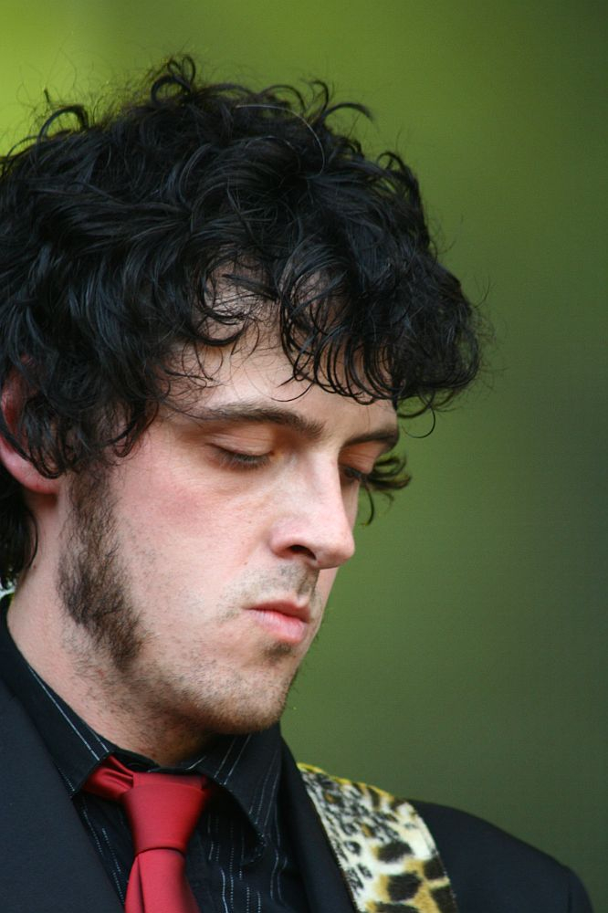 A man with curly dark hair, sideburns, and a pensive expression is shown looking down. Wearing a black jacket, red tie, and striped shirt, his attire suggests it might be from 2008. The background is blurred with a greenish tone, possibly indicating an outdoor setting at Positivus. The focus is on his face and upper body.