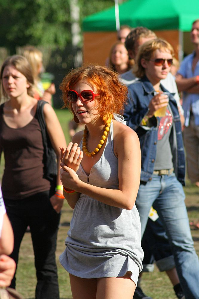 A young woman with bright orange hair, wearing red sunglasses, a grey dress, and a yellow beaded necklace, is walking outside among a crowd at Positivus 2008. Other people in casual attire are in the background, with some wearing jeans and T-shirts. Green and white tents are visible in the distance.