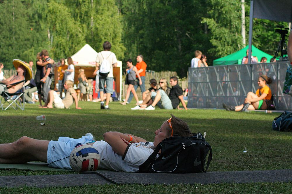 A boy wearing sunglasses lies on the grass next to a soccer ball and black backpack. In the background, people gather, sit, and socialize near white tents and a metal barrier at an outdoor event reminiscent of Positivus. Trees and greenery surround the open space, giving the scene a relaxed, summery atmosphere.