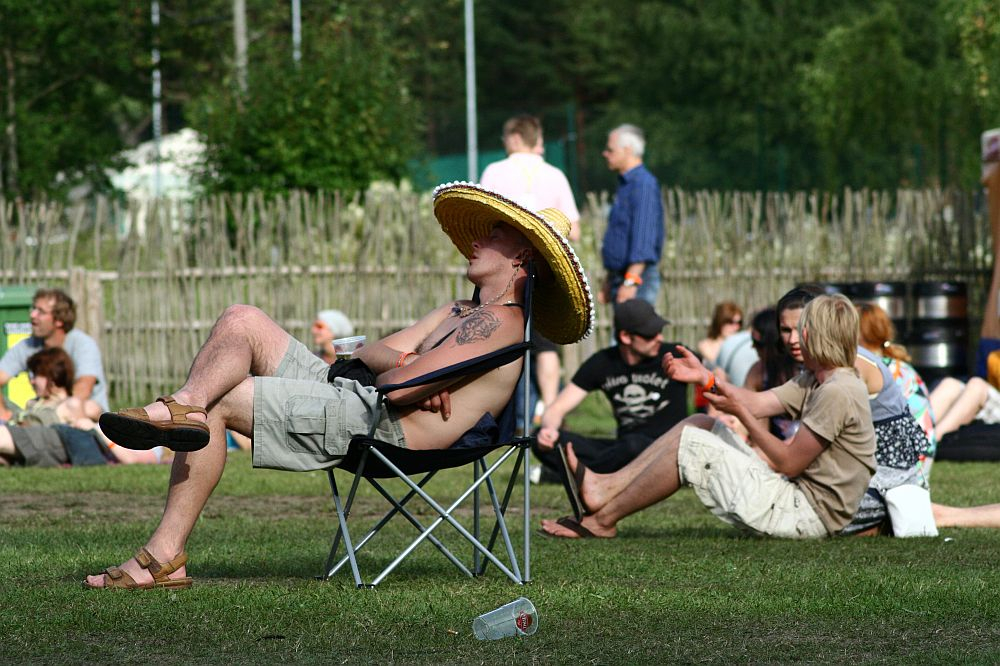 A man wearing a large sombrero and shorts reclines in a foldable chair on a grassy field during the 2008 Positivus festival, enjoying the lively outdoor event with other casually dressed attendees conversing in the background. A wooden fence and trees frame the distant AB landscape.