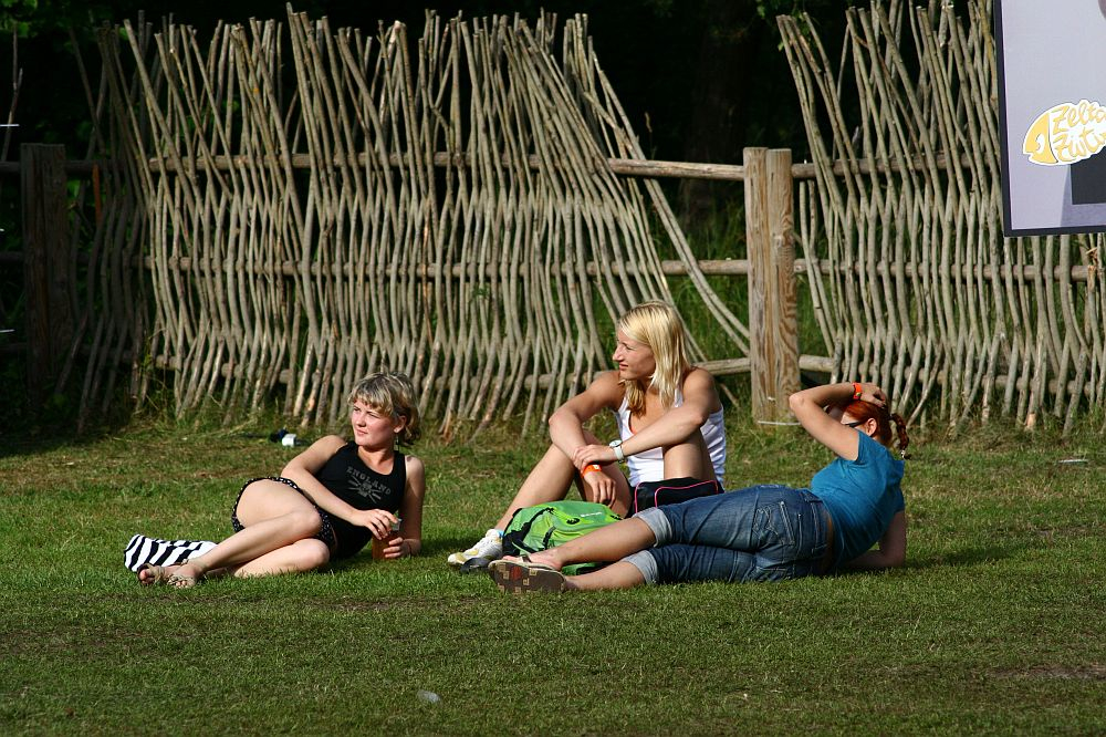Three people are sitting on grass near a wooden fence. Two are seated: one on the left in a black top and the other in the middle with a white top and green bag. A third person, wearing a blue top, is lying down on the right. The background channels Positivus 2008 vibes with its unique fence made of vertically interwoven branches.