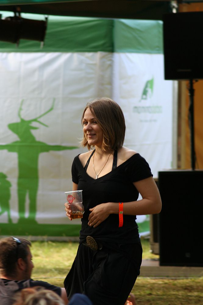 A smiling woman with shoulder-length brown hair wearing a black dress and an orange wristband holds a clear plastic cup. She stands under a green and white canopy with a shadowed deer emblem at Positivus AB 2008. People seated on the grass and sound equipment are visible in the foreground and background.