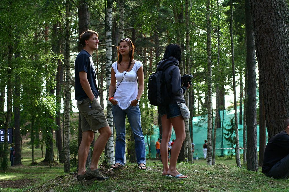 Three people stand in a wooded area with tall trees and dappled sunlight. Two women and a man appear to be having a conversation. One woman is holding a tablet, possibly referencing an event from 2008, while another is wearing a backpack. They are casually dressed, and the lush greenery suggests a park or forest setting akin to Positivus.