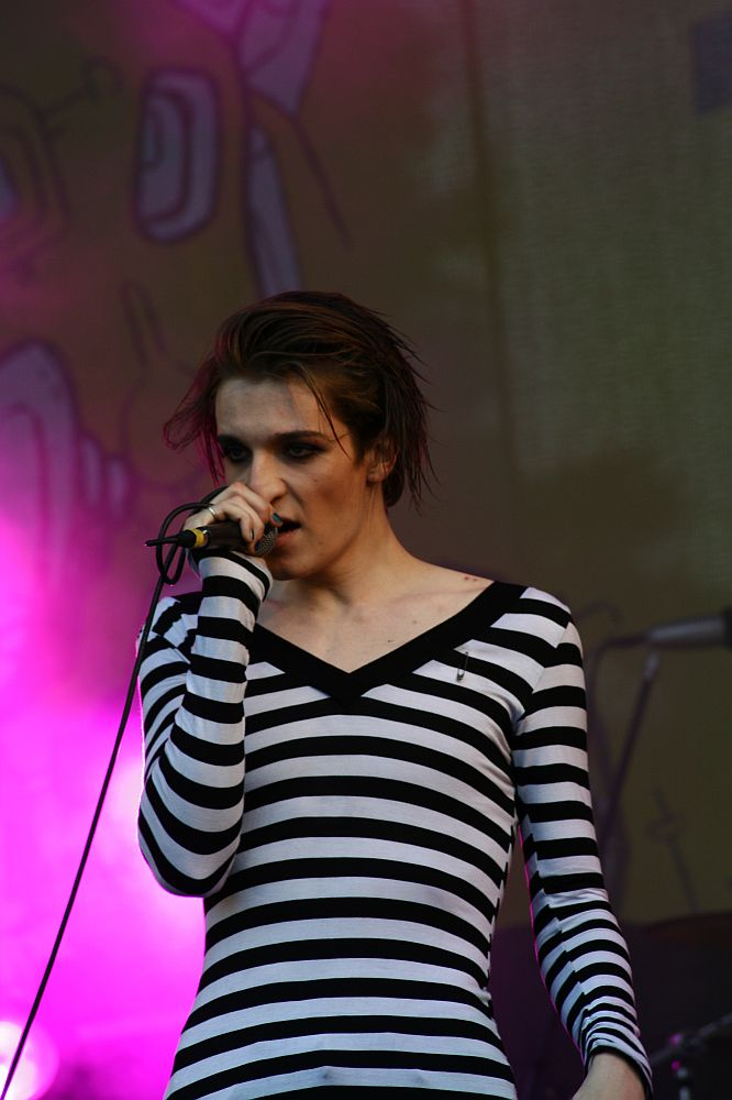 A person with short wet hair is performing on stage at Positivus, holding a microphone close to their mouth. They are wearing a black and white striped, long-sleeved top. The background is illuminated with vibrant purple and pink stage lights, reminiscent of the AB 2008 vibe. The overall atmosphere suggests a lively concert or music performance.