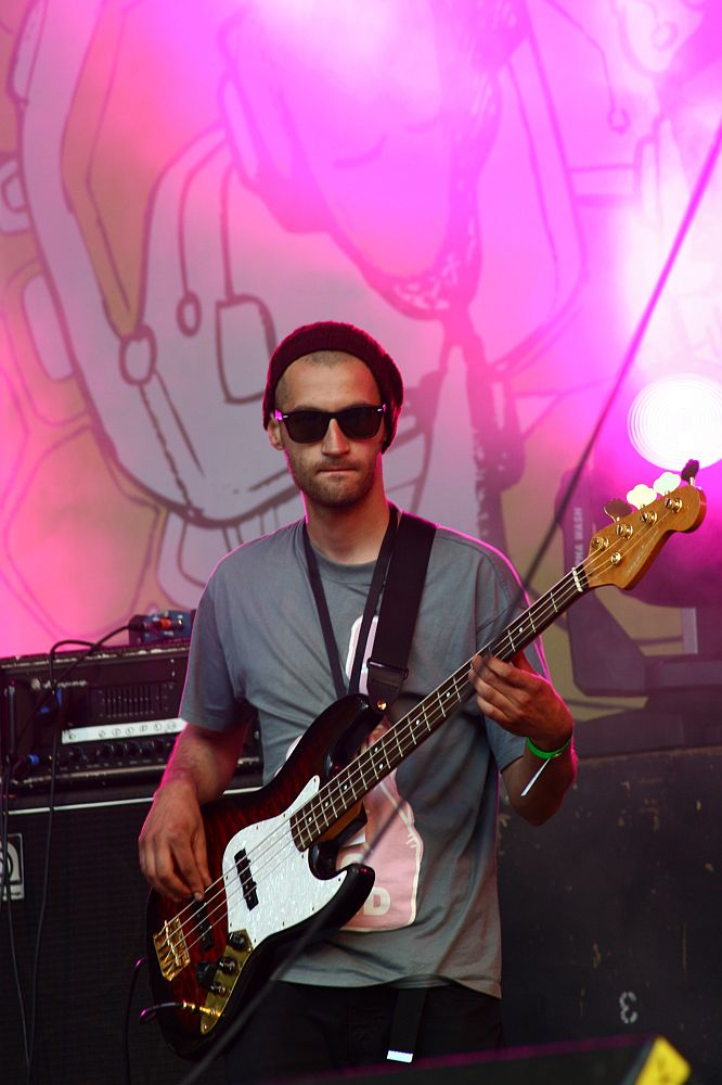 A musician wearing sunglasses, a beanie, and a T-shirt, playing an electric bass guitar on stage at the 2008 Positivus festival. The background features colorful, abstract artwork and pink stage lighting. He stands in front of AB audio equipment, immersed in his performance.